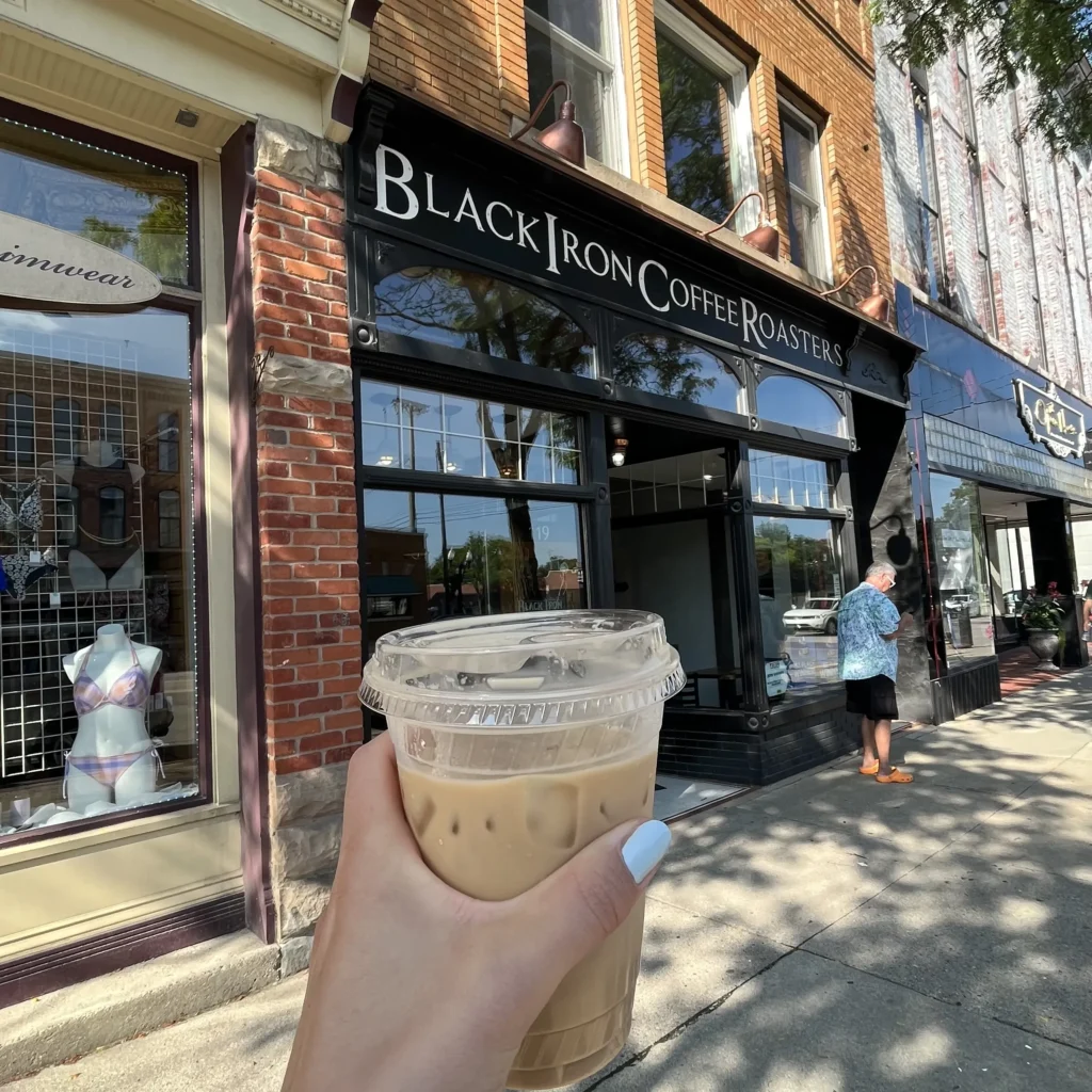 Hand holding iced latte in front of Black Iron Coffee Roasters store front.