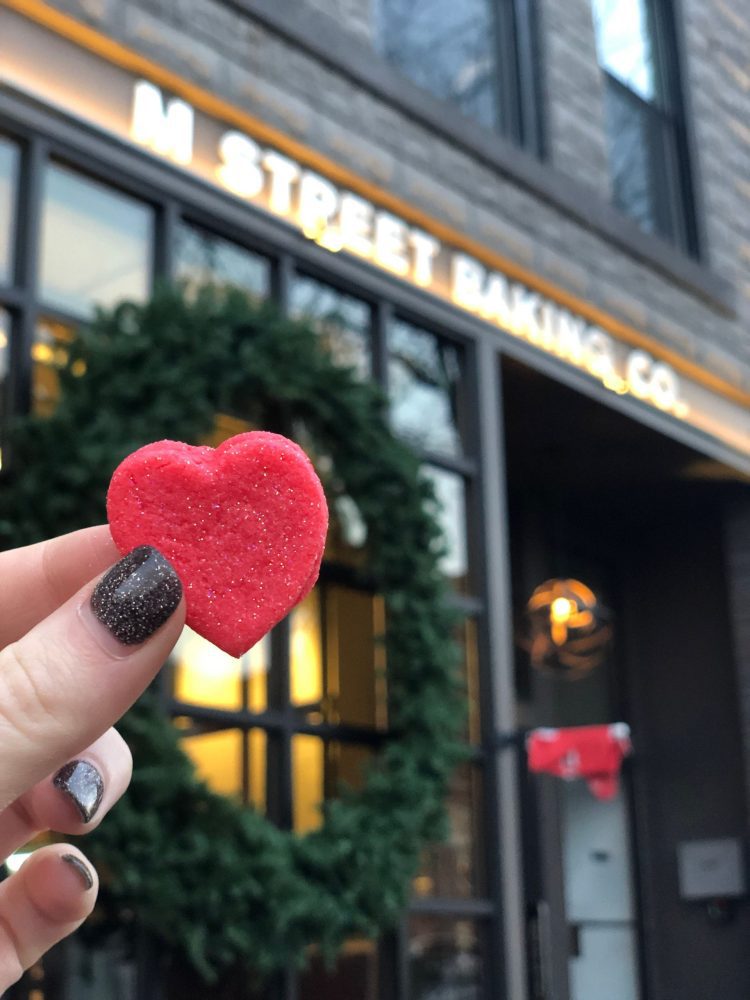 Hand holding a heart shaped cookie in front of M Street Baking Co