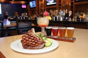 big seared meat decorated on the plate with a sample flight of beer and cocktail and bar behind.