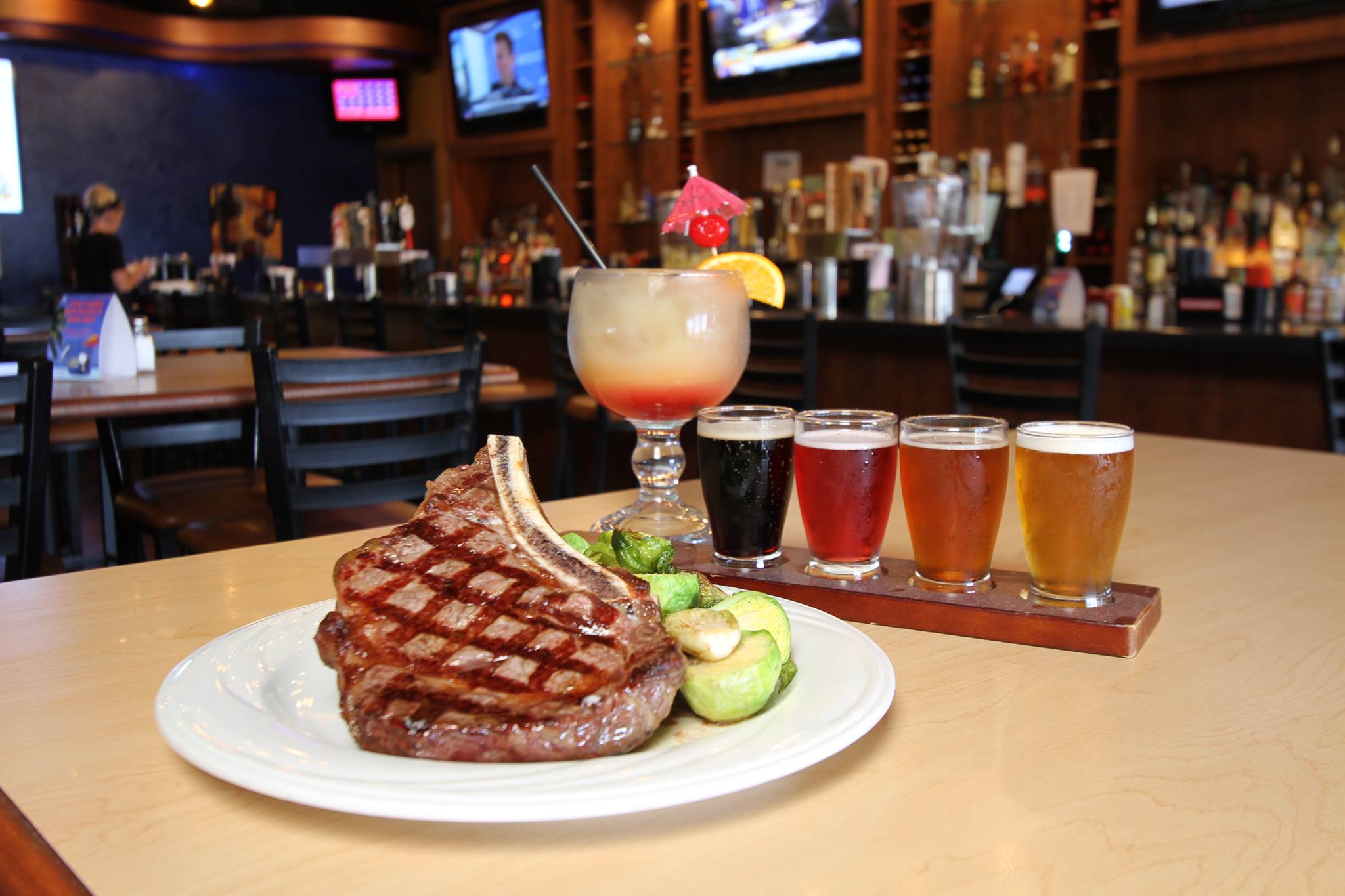 big seared meat decorated on the plate with a sample flight of beer and cocktail and bar behind.