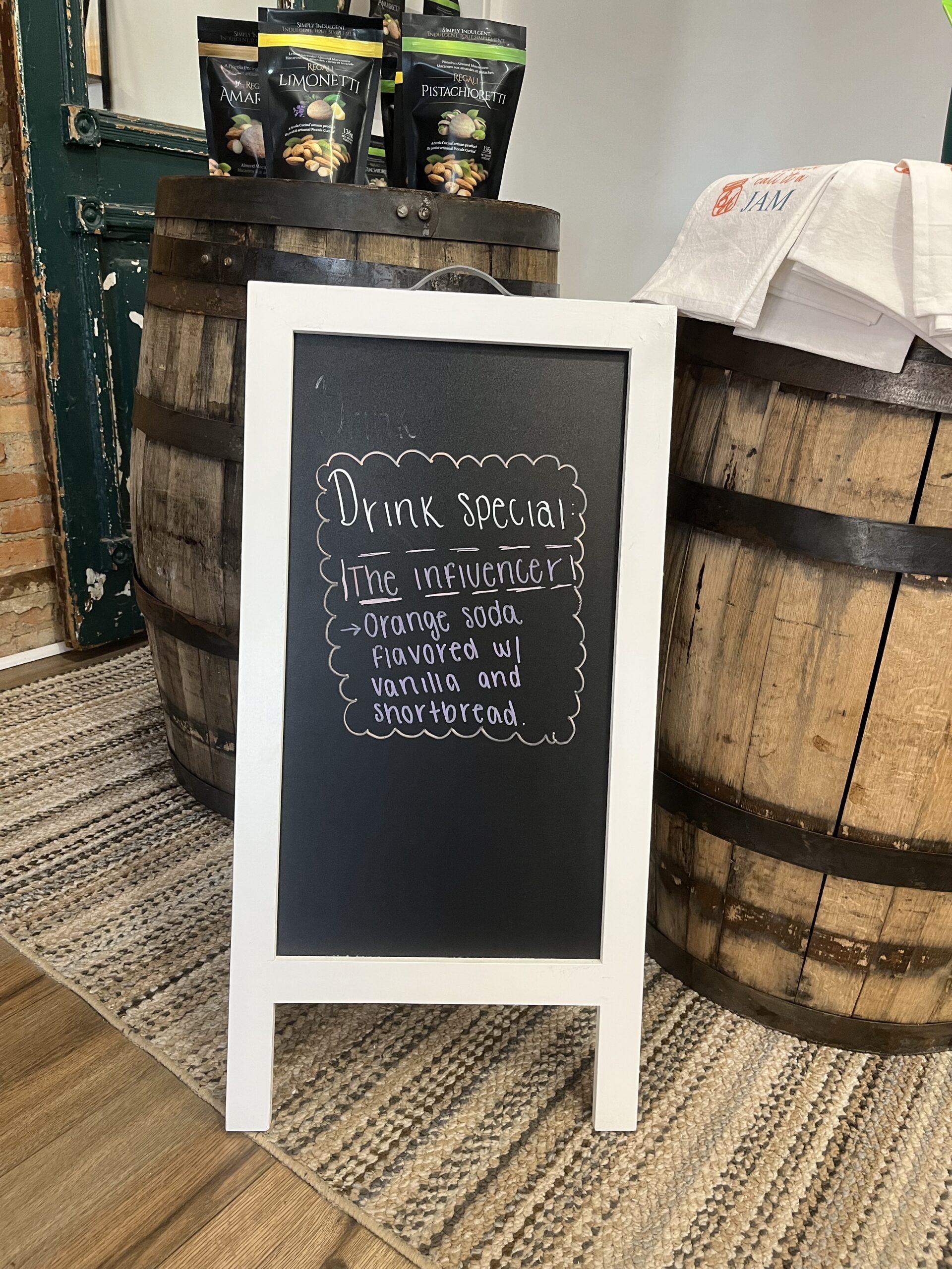 Drink specials board at Crunchy Pickle with barrels behind it selling merchandise. Chalk board with drink specials on rug.