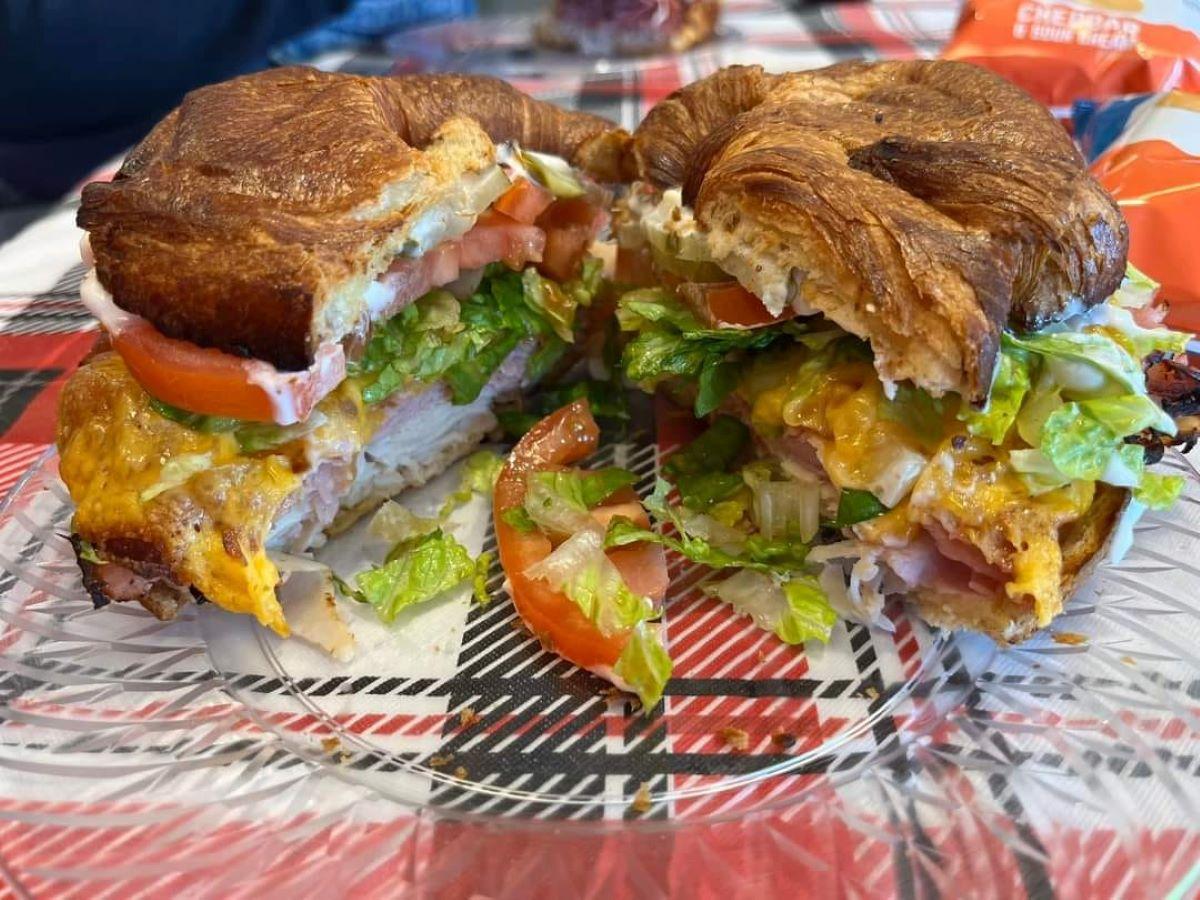 Burger sandwich servied on a croissant. Plated on plaid table cloth. Filled with red tomatos and green lettuce.